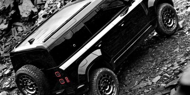 A 2026 Land Rover Defender in OCTA Black with a spare tire mounted on the rear drives up a rocky, uneven slope.