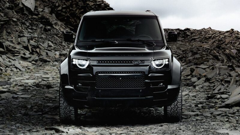 An OCTA Black Land Rover Defender is parked on a rocky terrain, viewed from the front with its headlights on under an overcast sky.