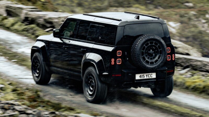 A 2026 Land Rover Defender in OCTA Black, with a spare tire mounted on the rear, drives on a wet, winding road through a rocky and grassy landscape.