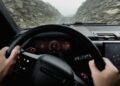 Navigating a narrow, rocky road in foggy weather, the driver grips the steering wheel of a 2026 Land Rover Defender OCTA Black, digital dashboard displays glowing through the mist.