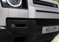 Close-up of the front bumper and grill of a white Land Rover Defender, highlighting the headlight, fog light, and license plate "143 UYP," showcasing recent design updates.