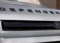 Close-up view of the front grille of a white Land Rover Defender, highlighting recent design updates with the bold "DEFENDER" lettering and the iconic Land Rover badge.