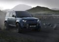 A blue Defender, featuring the latest design updates, drives on a rocky, dusty terrain with mountains and overcast skies in the background.