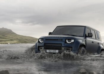 A blue Land Rover Defender showcasing its latest design updates drives through shallow water, set against a backdrop of mountains and cloudy skies.