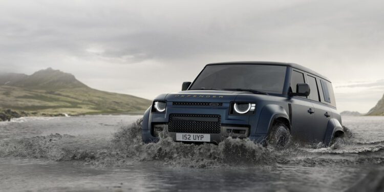 A blue Land Rover Defender showcasing its latest design updates drives through shallow water, set against a backdrop of mountains and cloudy skies.