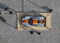 Overhead view of a silver race car with orange stripes displayed on a wooden platform at Luftgekhühlt 11, surrounded by people standing on gravel at the event location in Durham NC.