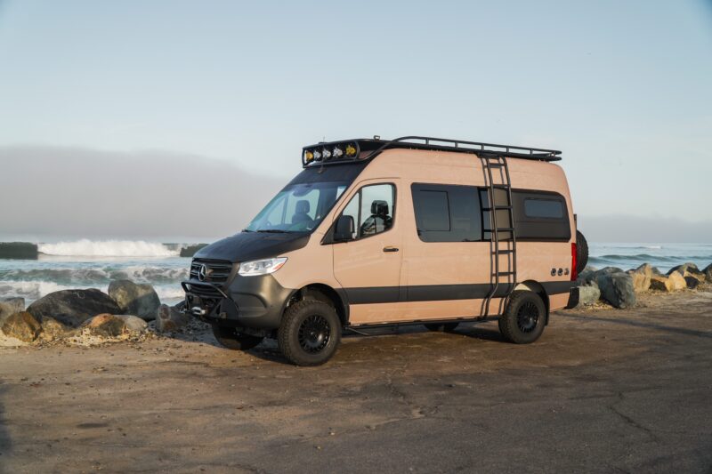 A tan and black Rogue Van Company camper van with off-road tires and a roof rack is parked by the ocean next to rocks under a clear sky.