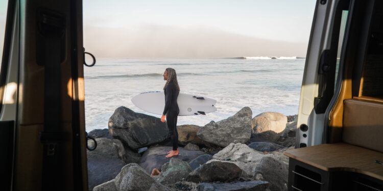 A person in a wetsuit stands barefoot on rocks by the ocean, holding a surfboard, viewed from inside a Rogue Van Company van.