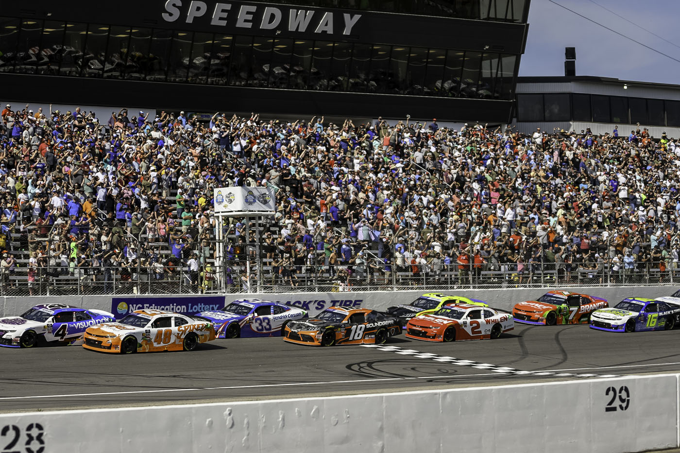 Race cars cross the starting line at Rockingham Speedway, the ultimate playground for racing fans, as a large crowd of spectators fills the grandstands under a sunny sky.