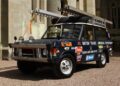 A black off-road vehicle, reminiscent of a Land Rover Classic to Showcase 10 Historic Expedition Icons at 2025 Pebble Beach Concours d'Elegance, is covered in expedition and sponsor stickers with a roof rack, spare tire on the hood, and a large ladder secured on top.