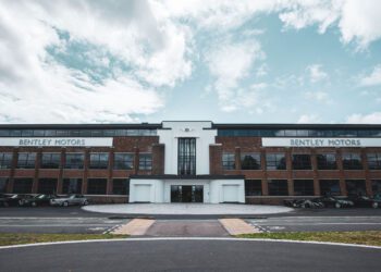 A large brick and glass building with "Bentley Motors" signs on both sides, several cars parked in front, and a partly cloudy sky above.