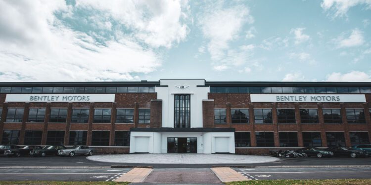 A large brick and glass building with "Bentley Motors" signs on both sides, several cars parked in front, and a partly cloudy sky above.