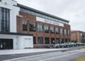 Brick and white Bentley design studio with large windows, sign above entrance, and several Bentley cars parked in front on a cloudy day.