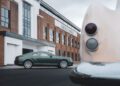 A green Bentley coupe is parked outside a brick Bentley Motors design studio, with part of a classic car’s rear in the foreground.