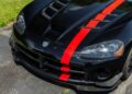 Close-up of the front end of a black sports car with a red racing stripe, parked on a driveway next to grass. Bid On This 2010 Dodge Viper SRT-10 Roadster ACR With Just 20 Miles for an iconic addition to your collection.