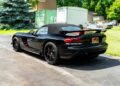 A black convertible sports car, possibly like the 2010 Dodge Viper SRT-10 Roadster ACR with just 20 miles you can bid on, is parked on a paved driveway near grass, trees, and shipping containers.