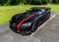 A black convertible sports car with red racing stripes, this 2010 Dodge Viper SRT-10 Roadster ACR with just 20 miles is parked on a driveway beside a grassy area and trees. Bid On This rare find today!.