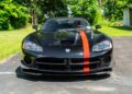 Black sports car with a prominent red racing stripe parked on a driveway, surrounded by green grass and trees in the background. Bid On This 2010 Dodge Viper SRT-10 Roadster ACR With Just 20 Miles and make it yours today.
