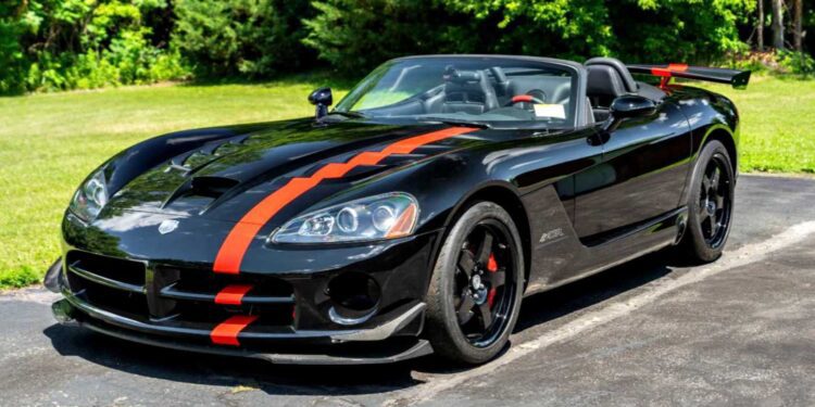 A black convertible sports car with red racing stripes and a rear spoiler is parked on a paved driveway with greenery in the background. Bid On This 2010 Dodge Viper SRT-10 Roadster ACR With Just 20 Miles today!.