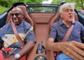 Two men are seated in a convertible, both wearing seatbelts; the man on the right is driving as they discuss The W16 Swan Song: Jay Leno Shares His Thoughts On The Bugatti Mistral while cruising down a green, tree-lined road.