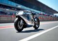 A sleek, silver motorcycle—rideable art—rests on a racetrack pit lane, with blurred figures and a grandstand in the background.