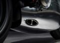 Close-up of a polished metal vehicle body, designed as a futuristic aluminum sculpture, with a dual exhaust outlet near the rear wheel reflecting light and surrounding surfaces.
