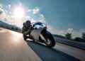 A motorcyclist leans into a curve on a racetrack at high speed, riding a limited edition sculpture that blurs the line between rideable art and machine, with sunlight and mountains in the background.