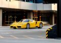 A bright yellow Ferrari Enzo in Giallo Modena is parked in an empty, modern urban parking area with reflective glass buildings in the background.