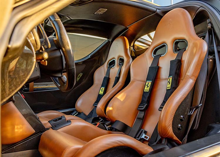 Close-up view of a luxury Ferrari Enzo interior featuring two Cuoio Leather racing seats with harness seatbelts and visible Ferrari logos.