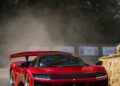 A red Ferrari sports car with a rear wing speeds around a corner at Goodwood, kicking up dust past hay bales, with trees visible in the background.