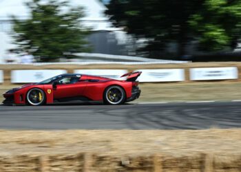 A red Ferrari speeds along the Goodwood racetrack, bordered by hay bales, with trees and white fencing in the background.