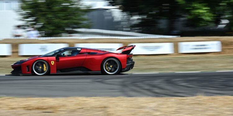 A red Ferrari speeds along the Goodwood racetrack, bordered by hay bales, with trees and white fencing in the background.