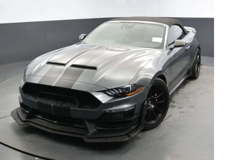 A gray convertible sports car with black wheels is parked indoors on a concrete floor in a garage, facing slightly to the left.