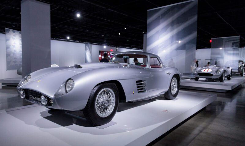A silver classic sports car is displayed on a platform in a modern, well-lit automobile museum, with another vintage car visible in the background.