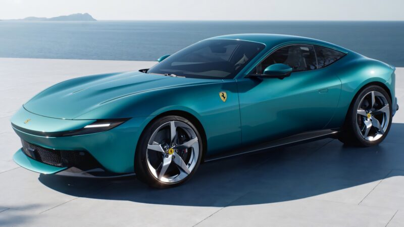 A teal Ferrari Amalfi sports car is parked on a light-colored surface near an ocean, with distant islands visible in the background.