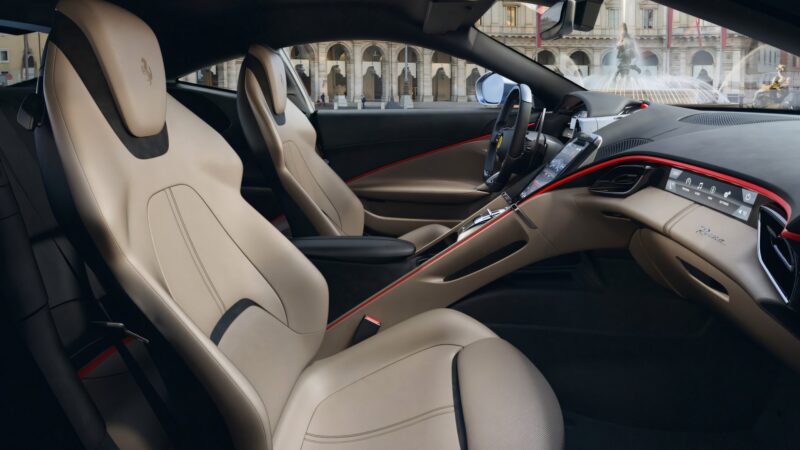 The image shows the beige leather interior of a modern Ferrari Roma sports car with two seats, a digital dashboard, and city buildings visible through the window.