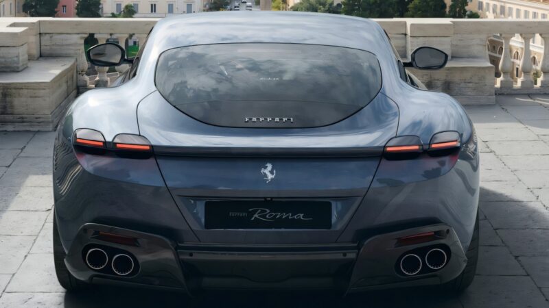 A rear view of a grey Ferrari Roma Facelift parked outdoors, showcasing its taillights, quad exhaust pipes, and badge.