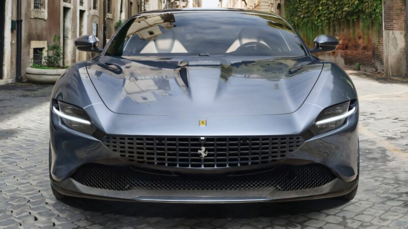A silver Ferrari Roma Facelift is parked on a cobblestone street between old buildings, viewed from the front.