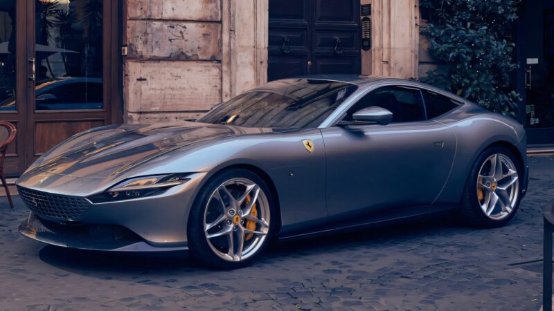 A silver Ferrari Roma Facelift sports car is parked on a cobblestone street in front of a building with stone walls and lush plants.
