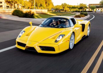 A yellow Ferrari Enzo in striking Giallo Modena, featuring luxurious Cuoio Leather seats, cruises down a multi-lane road lined with trees and buildings on a sunny day with two people inside.