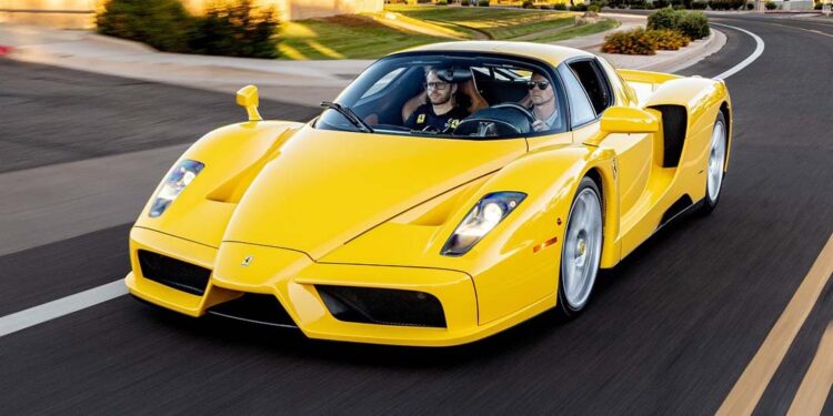 A yellow Ferrari Enzo in striking Giallo Modena, featuring luxurious Cuoio Leather seats, cruises down a multi-lane road lined with trees and buildings on a sunny day with two people inside.