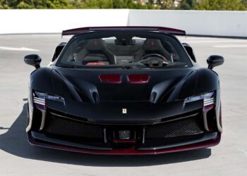 Front view of a black Ferrari Beverly Hills SF90 XX Spider with striking red carbon fiber accents, parked on a rooftop against trees and a white barrier backdrop.
