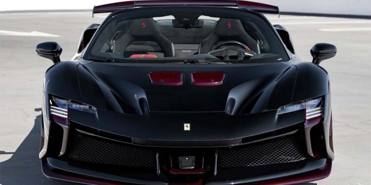 Front view of a black Ferrari Beverly Hills SF90 XX Spider with striking red carbon fiber accents, parked on a rooftop against trees and a white barrier backdrop.