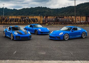 Three blue Porsche sports cars, including a Porsche 991 GT2 RS and a GT3 Touring, are parked on asphalt in front of a freight train with trees and hills in the background.