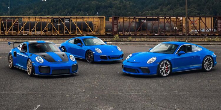 Three blue Porsche sports cars, including a Porsche 991 GT2 RS and a GT3 Touring, are parked on asphalt in front of a freight train with trees and hills in the background.