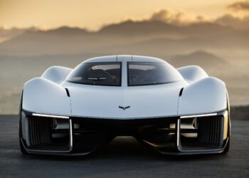 A futuristic white Corvette, an American icon, is parked outdoors facing the camera, set against mountains and a hazy sunset sky.
