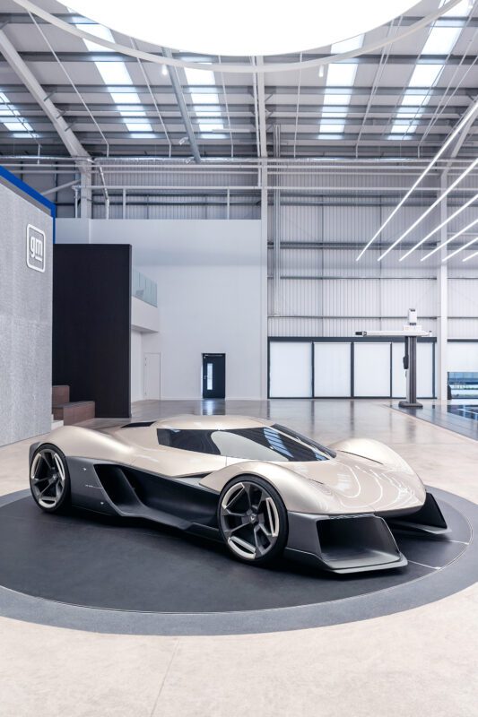 A futuristic silver Corvette concept car with a low, aerodynamic design is displayed on a circular platform inside a modern, spacious showroom, embodying future design and the spirit of an American icon.