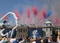 Crowds watch planes perform an aerial show with red, white, and blue smoke above a historic building during an outdoor event celebrating the 10 Fastest Cars At The 2025 Goodwood Festival Of Speed.