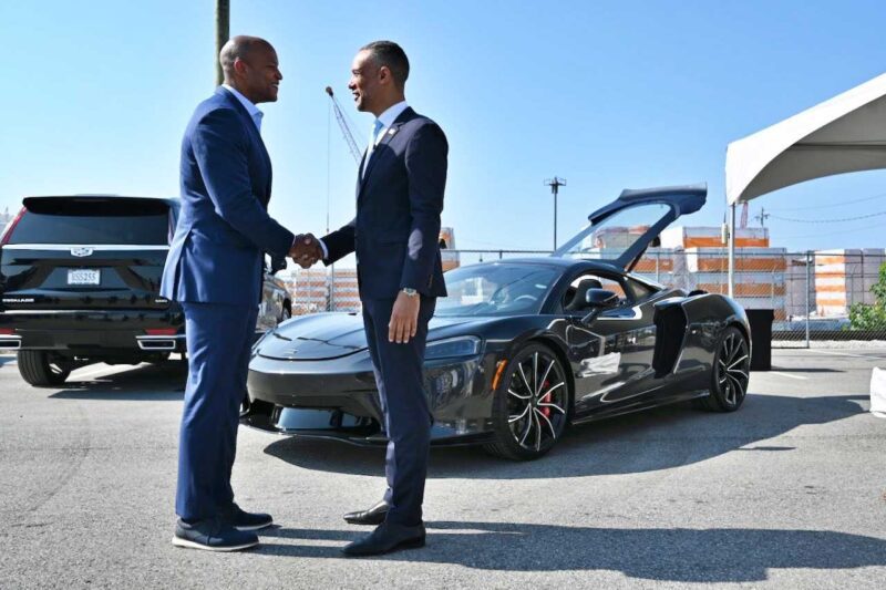 Two men in suits shake hands in front of a black sports car with its door open, outdoors on a sunny day—celebrating as McLaren doubles down on America, breaking ground on a $10.5m Vehicle Processing Center in Baltimore.