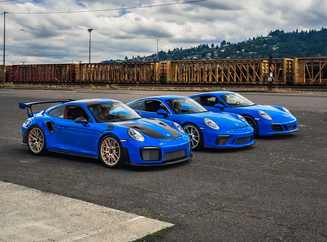 Three blue Porsche sports cars, including a GT3 and a Porsche 991, are parked side by side on an asphalt surface near a train yard with freight cars in the background.